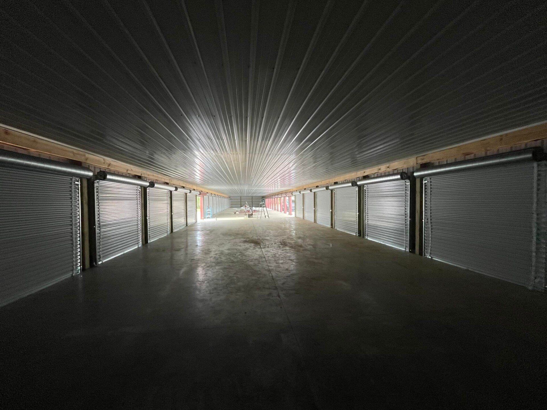 Interior of a storage facility with rows of closed metal roll-up doors, concrete floor, and metallic ceiling.