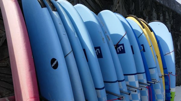 Hilera de coloridas tablas de surf apiladas verticalmente al aire libre, en su mayoría azules con tablas rosas y amarillas.