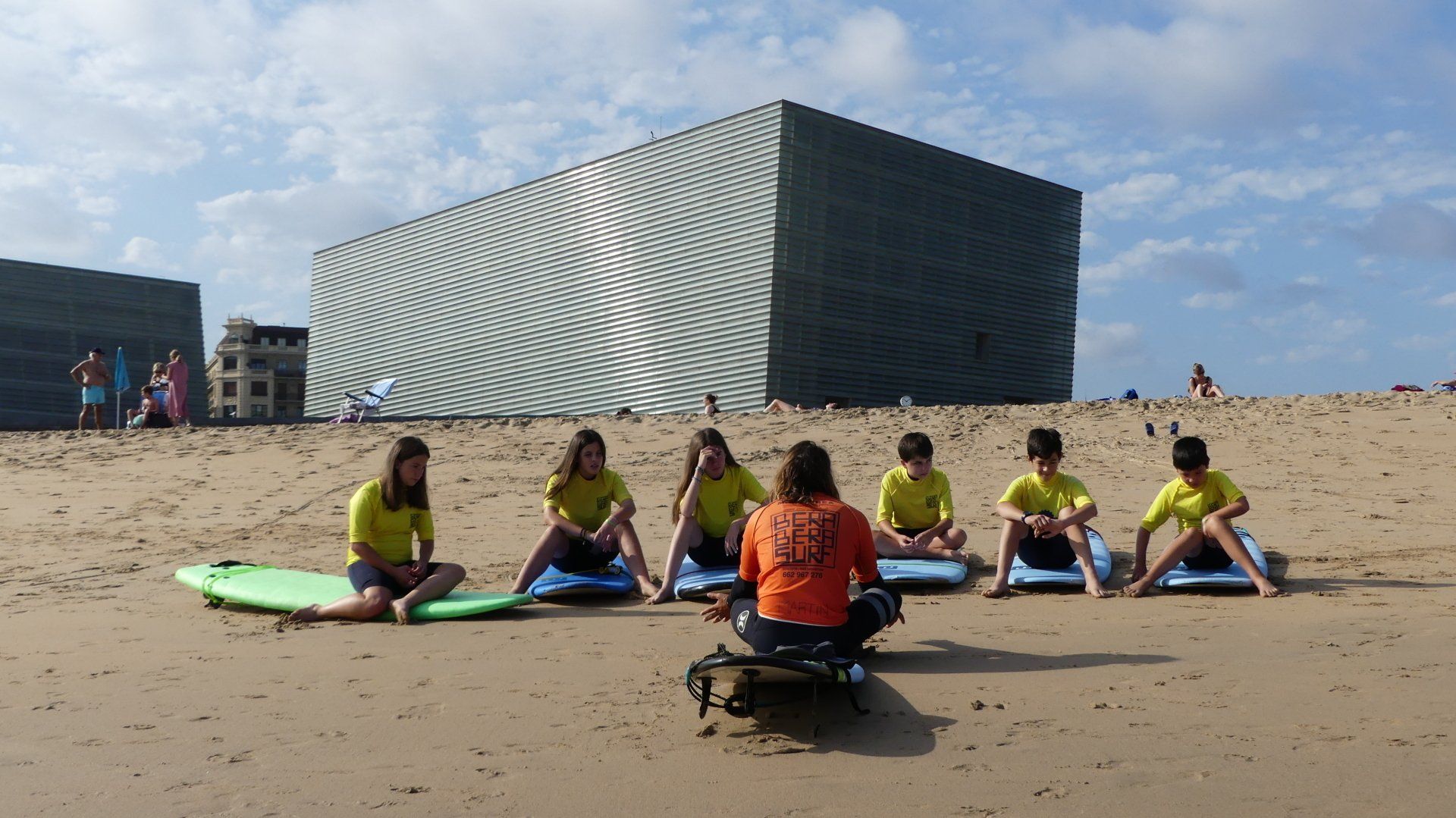 Clase de surf en una playa de arena con un grupo sentado en tablas frente a un instructor, con un edificio moderno al fondo.