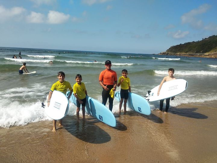 Surfistas con tablas blancas en una playa soleada con olas pequeñas y una costa rocosa.