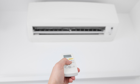 A hand holds a white remote control pointed toward a white wall-mounted air conditioner.