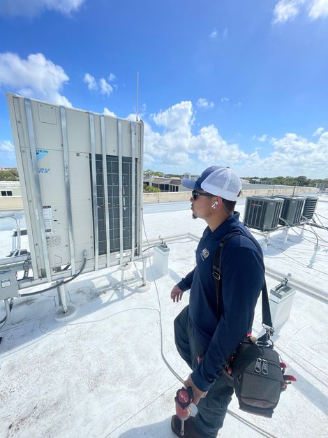 A technician in a navy shirt and cap stands on a white rooftop, examining industrial HVAC equipment.