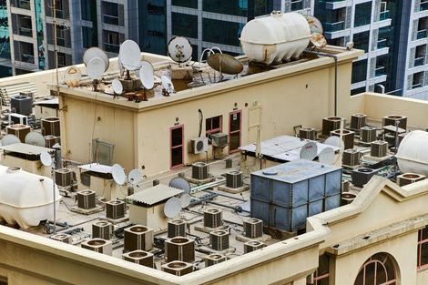 A high-angle view of a beige building rooftop cluttered with numerous satellite dishes, HVAC units, and water tanks.