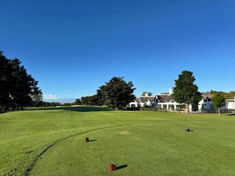 A sunny golf course tee box with a green fairway, scattered trees, and white buildings in the background.