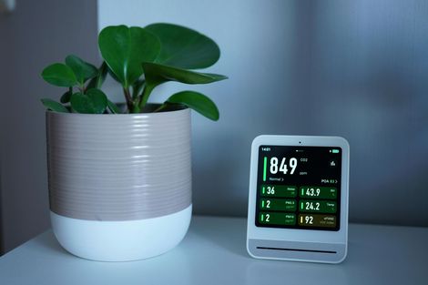 A small, digital air quality monitor displaying data next to a potted green plant on a white surface.