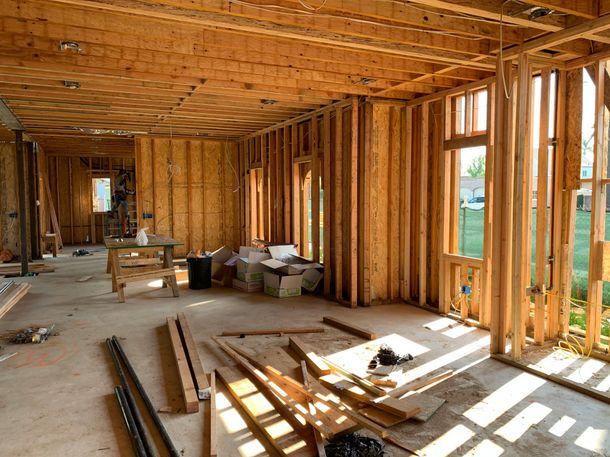 Interior view of a house under construction with exposed wooden wall studs, floor joists, and scattered building materials.