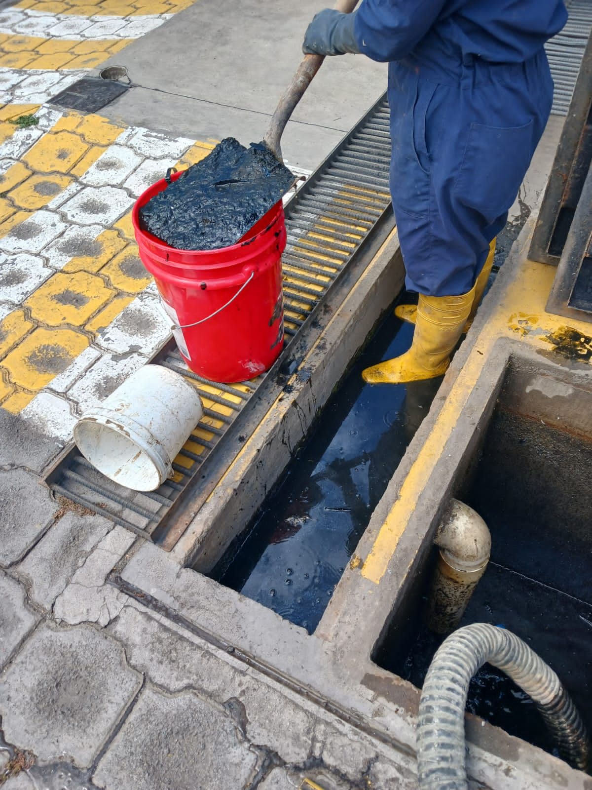 Trabajador con mono azul y botas amarillas limpiando una alcantarilla con un cubo y una pala rojos.