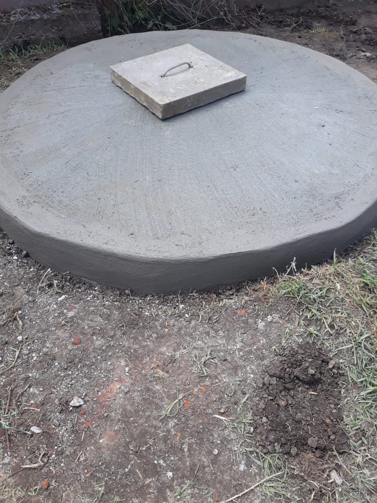 Tapa de alcantarilla redonda de hormigón con una pequeña trampilla de acceso cuadrada en la parte superior, colocada sobre tierra y césped.