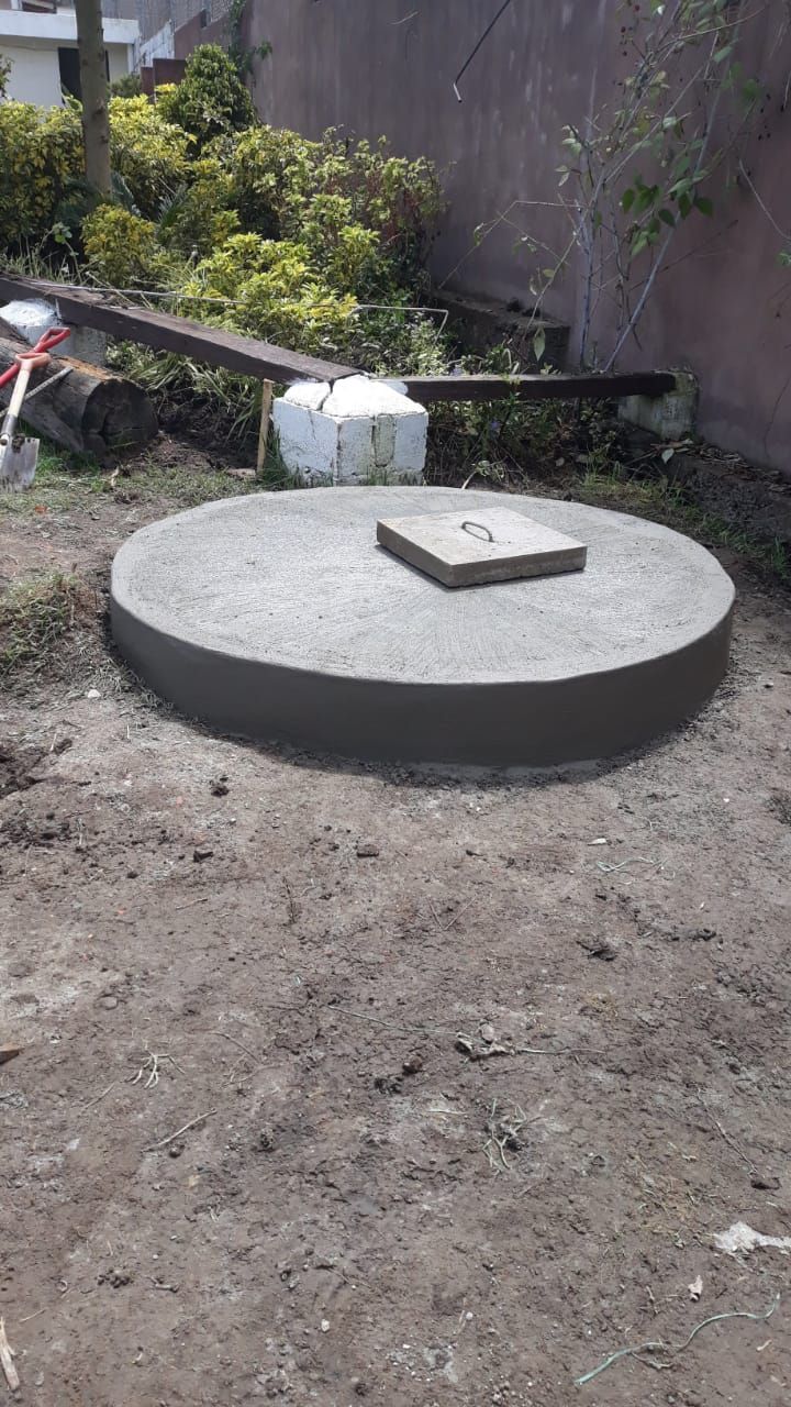 Plataforma circular de hormigón en un patio de tierra con un pequeño bloque rectangular encima, junto a un muro y plantas.