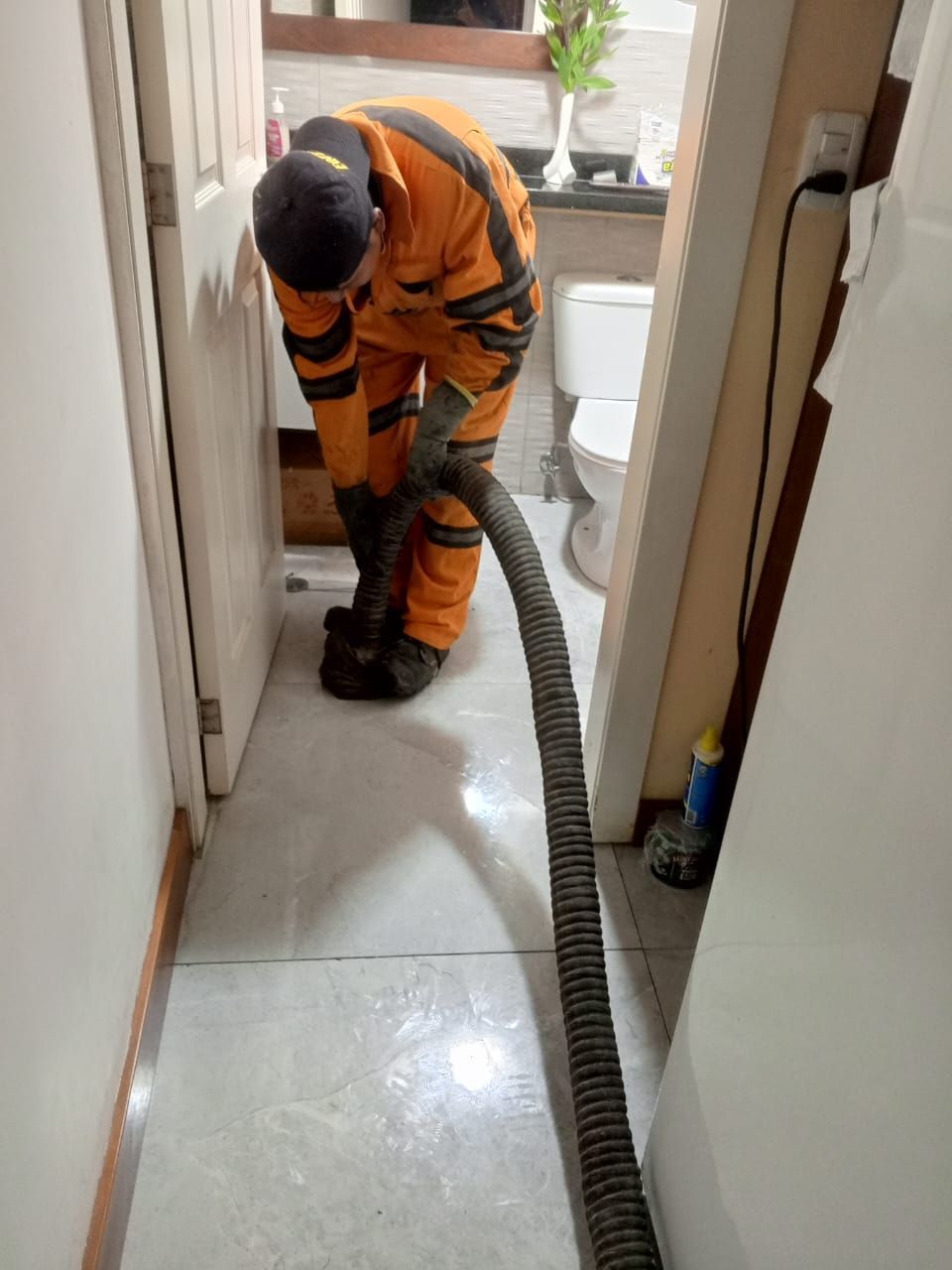 Un trabajador con un mono naranja aspira el suelo de un pasillo cerca de un baño.