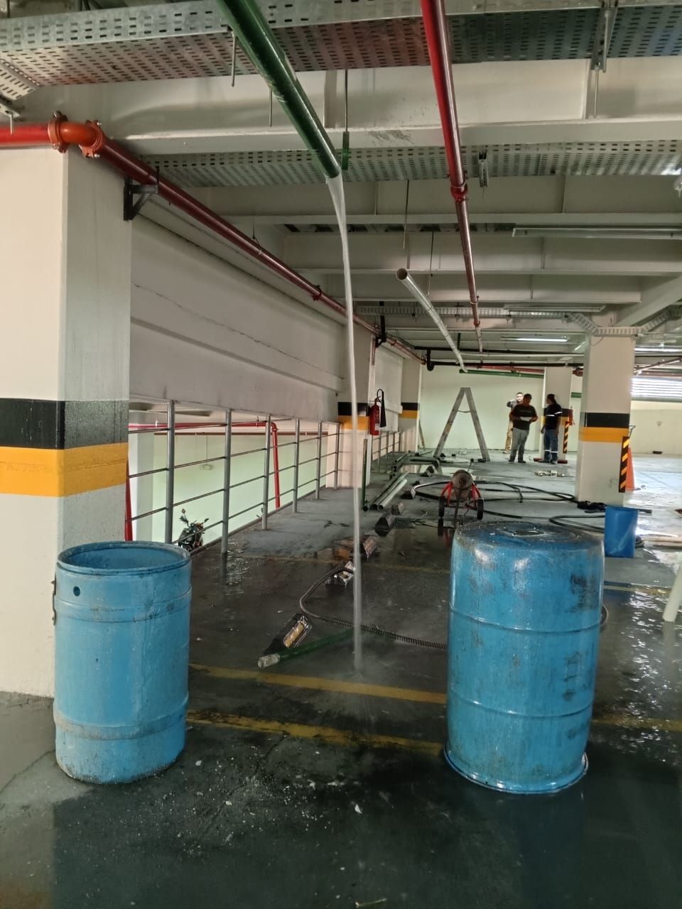 Garaje industrial con barriles azules, tuberías rojas y suelo mojado bajo una pasarela cubierta.