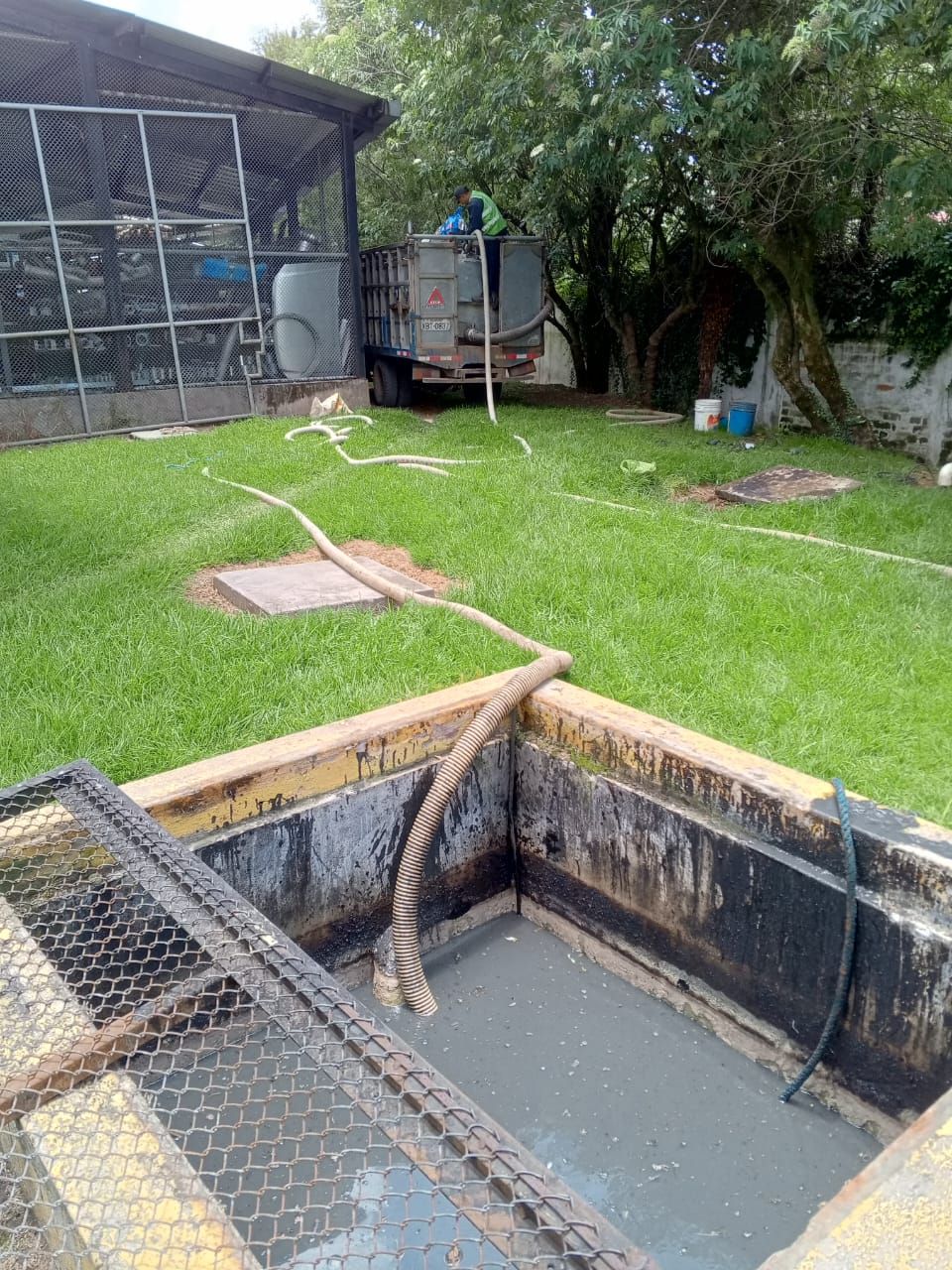 Pozo de servicios en el patio trasero con trampilla abierta, cubierta de malla y una manguera sobre césped verde cerca de un área cercada.