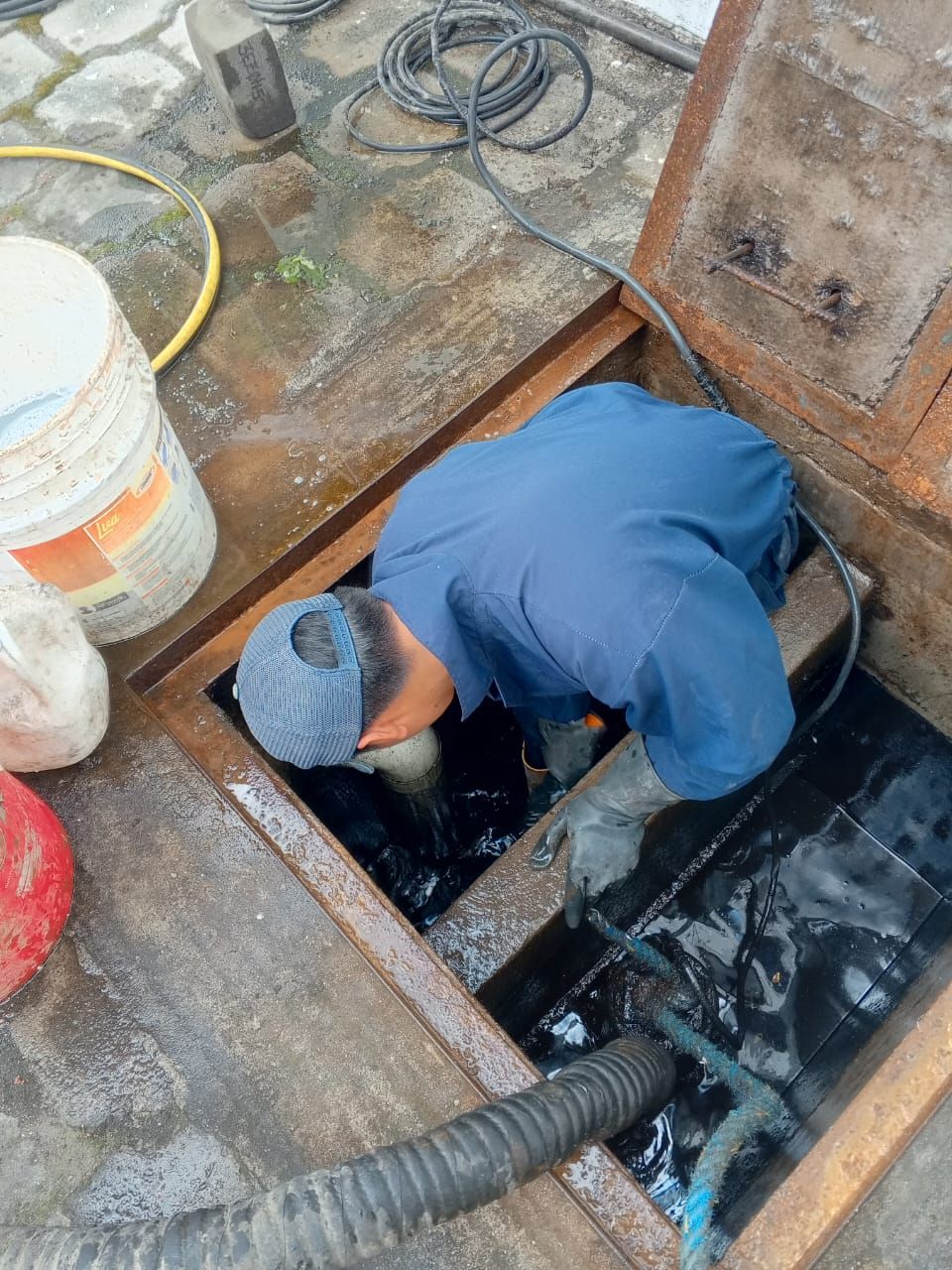 Un trabajador vestido de azul limpia un pozo negro dentro de una abertura en el piso de concreto, cerca de cubos y mangueras.