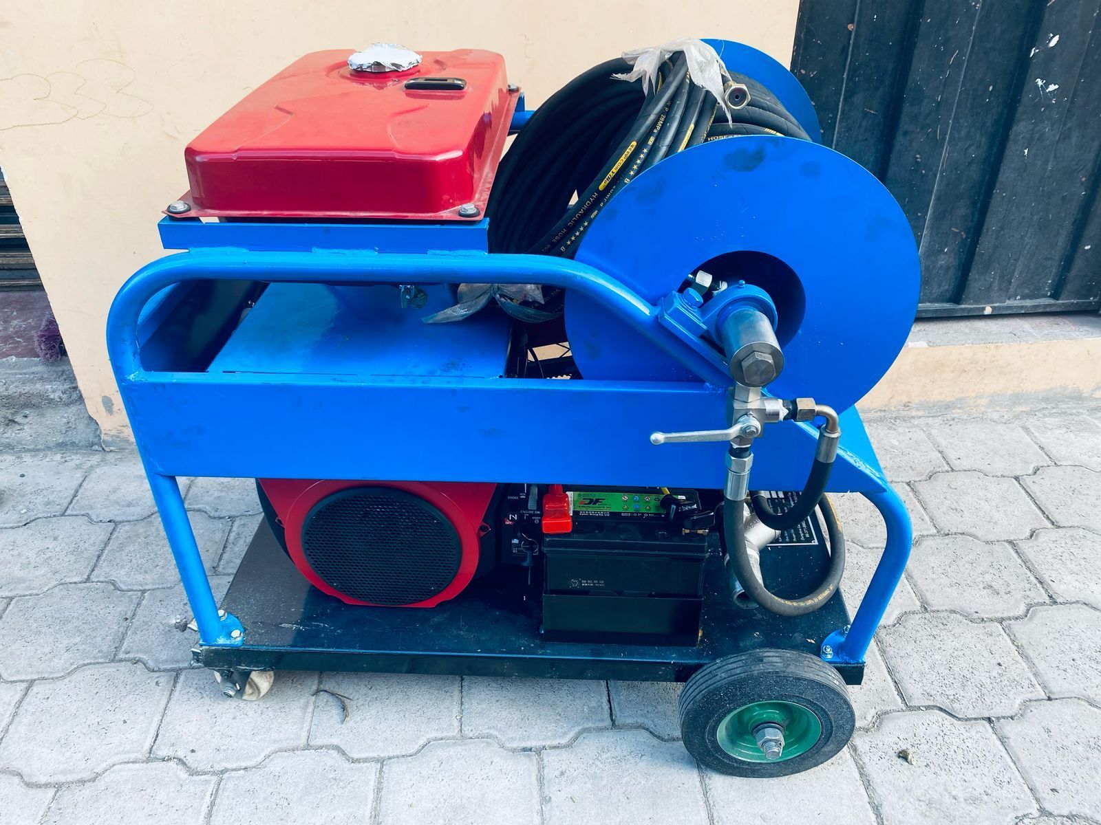 Máquina con estructura azul, depósito de combustible rojo y rueda azul grande sobre una base con ruedas, al aire libre.