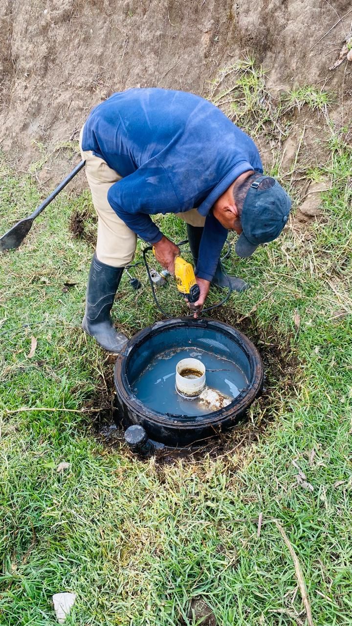 Un trabajador con chaqueta azul se inclina sobre un pozo abierto, utilizando una herramienta para inspeccionarlo o repararlo.