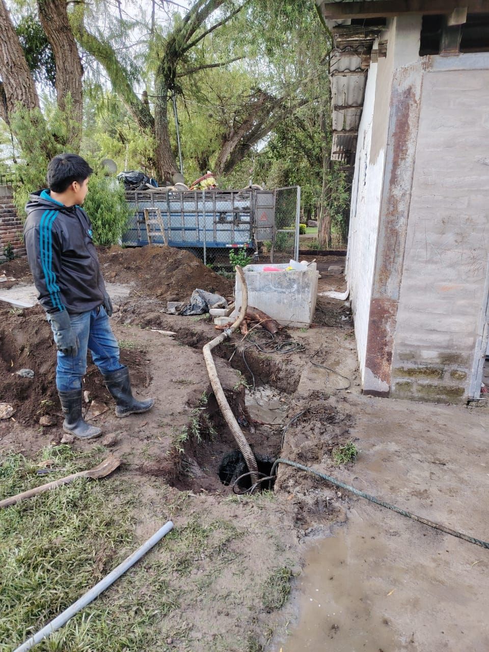 Una persona está de pie junto a una zanja fangosa y una tubería expuesta al lado de un pequeño edificio en un patio.