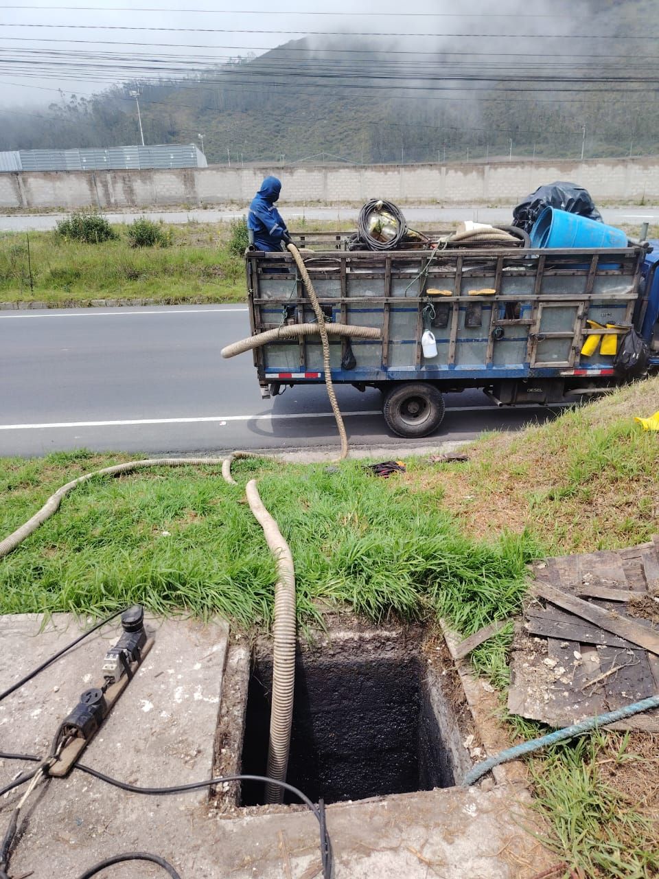 Un trabajador en un camión de servicios públicos junto a una alcantarilla abierta al borde de la carretera, cerca de una zanja cubierta de hierba.
