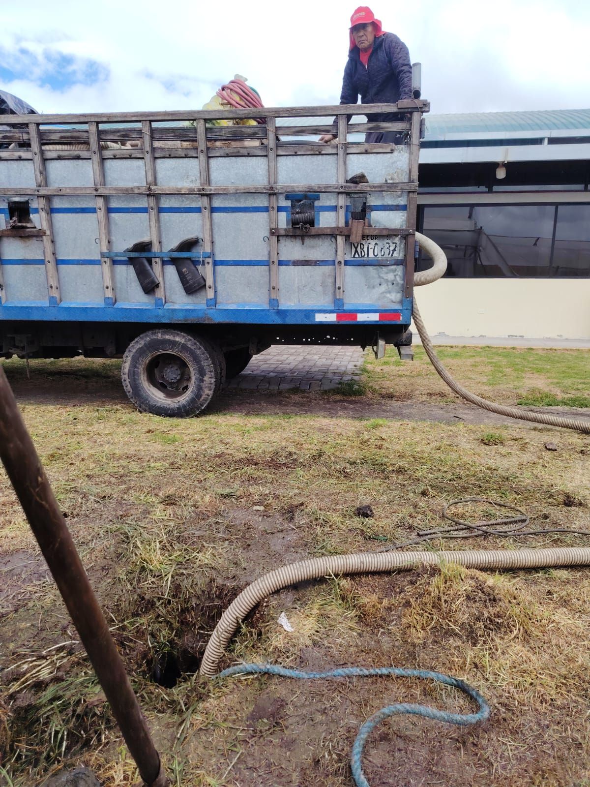 Un trabajador en la caja de una camioneta azul rocía con una manguera un campo embarrado al aire libre.