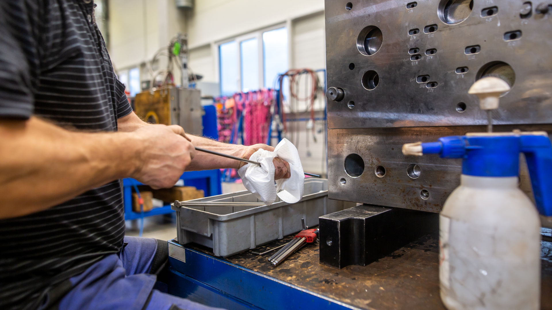 Worker using a power tool on metal parts at a workshop bench with a blue spray bottle nearby