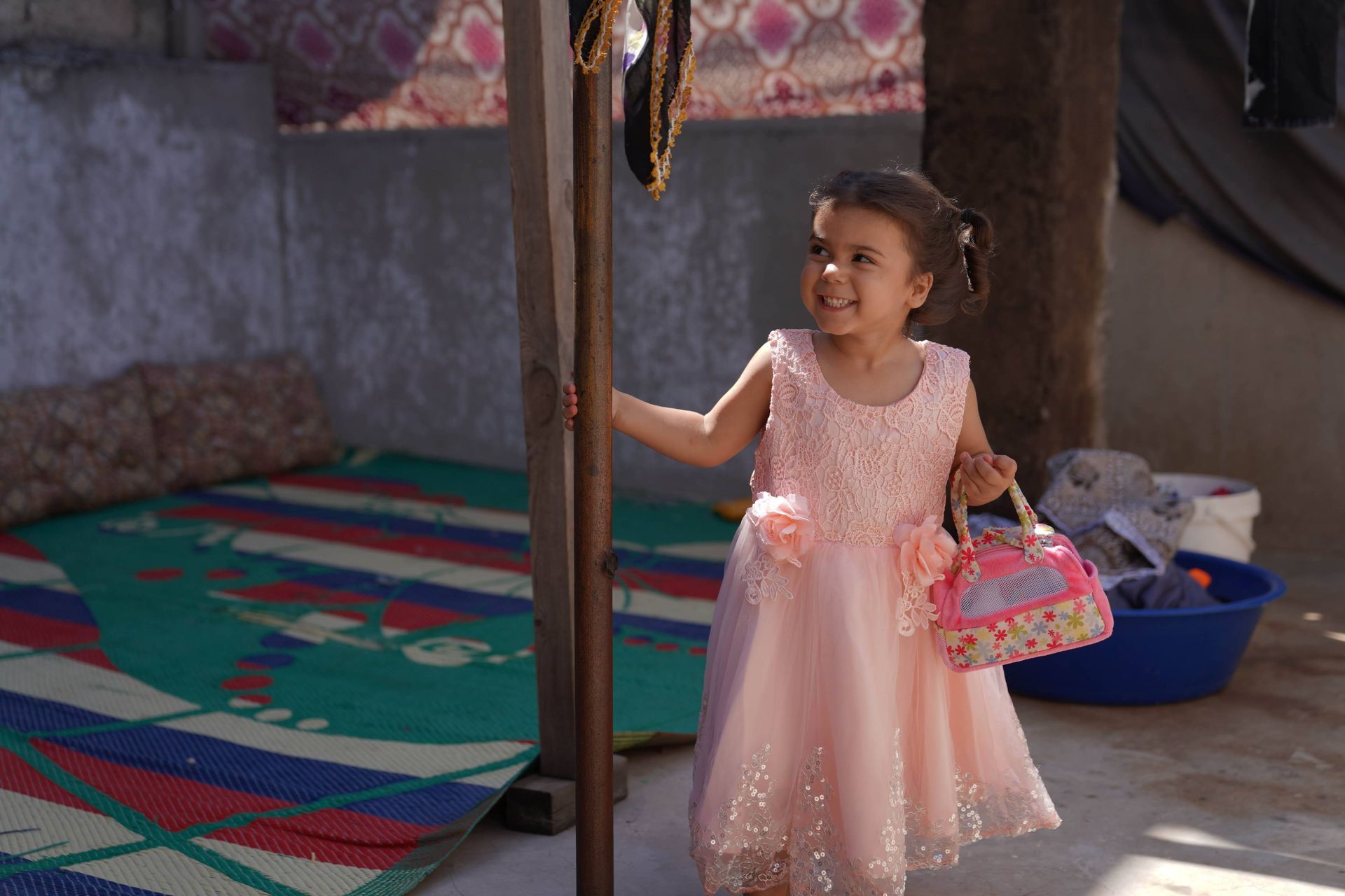 Une enfant souriante, vêtue d'une robe rose pâle, se tient près d'un poteau en bois, tenant un panier décoratif coloré.