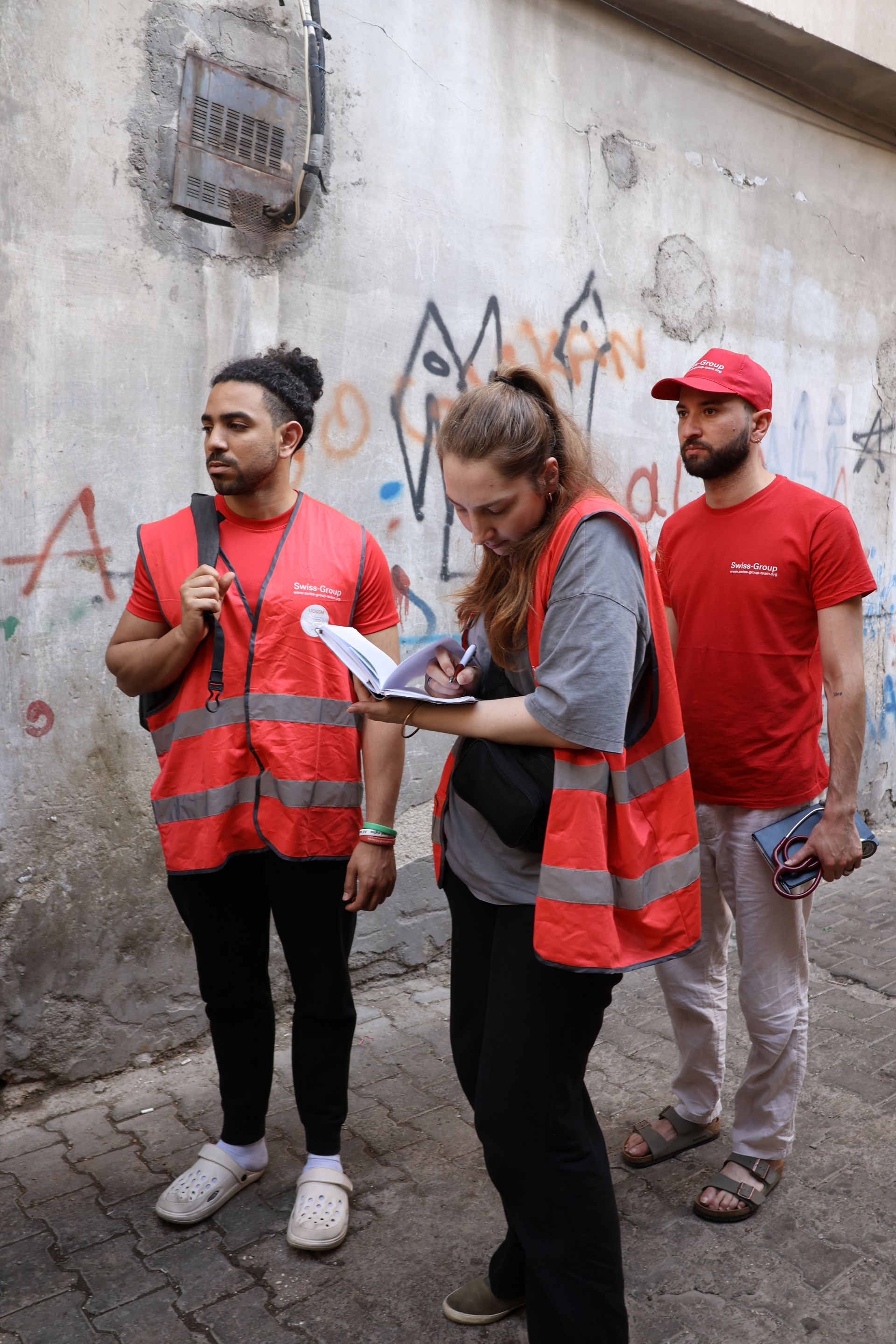 Trois individus vêtus de gilets et de chemises de sécurité rouges se tiennent dans la rue, celui du milieu écrivant dans un carnet.