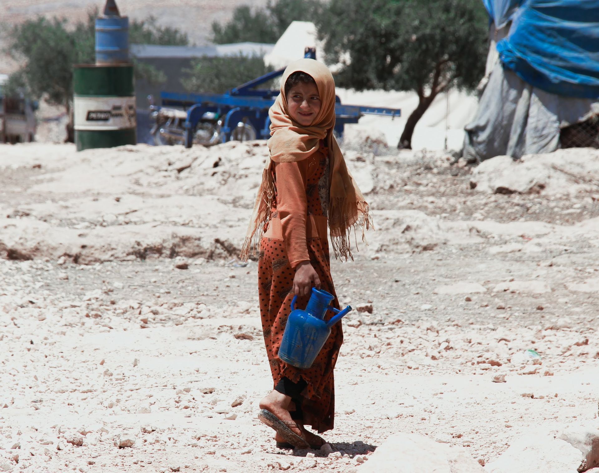 Une personne vêtue d'une robe à motifs et d'un foulard traverse une zone rocailleuse et aride en portant un récipient d'eau en plastique bleu.