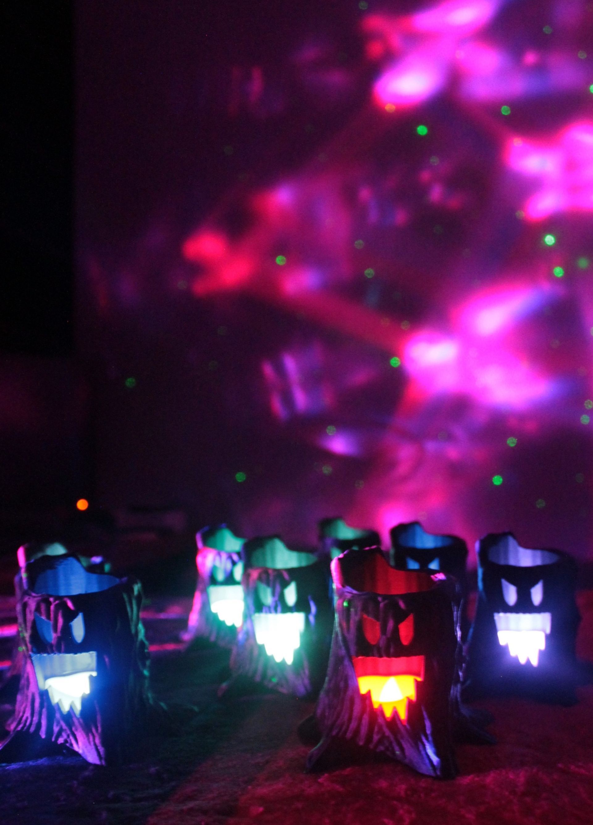 Lit-up ghost tree decorations with colorful glowing eyes in front of a purple and green nebula backdrop.