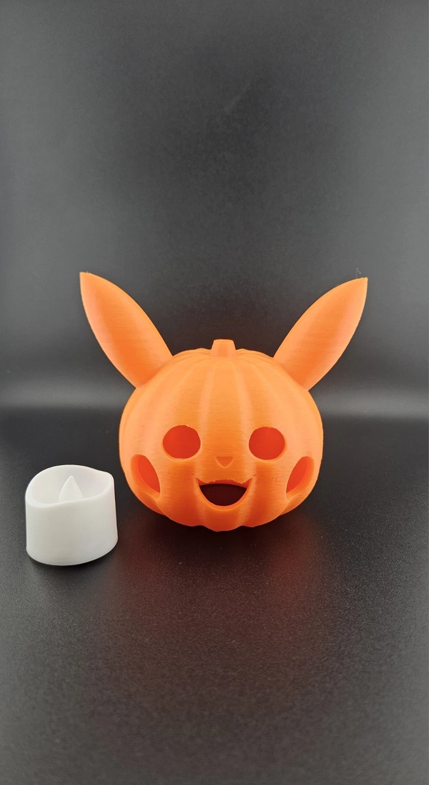 Orange 3D-printed pumpkin pikachu and a white tea light on a black surface.