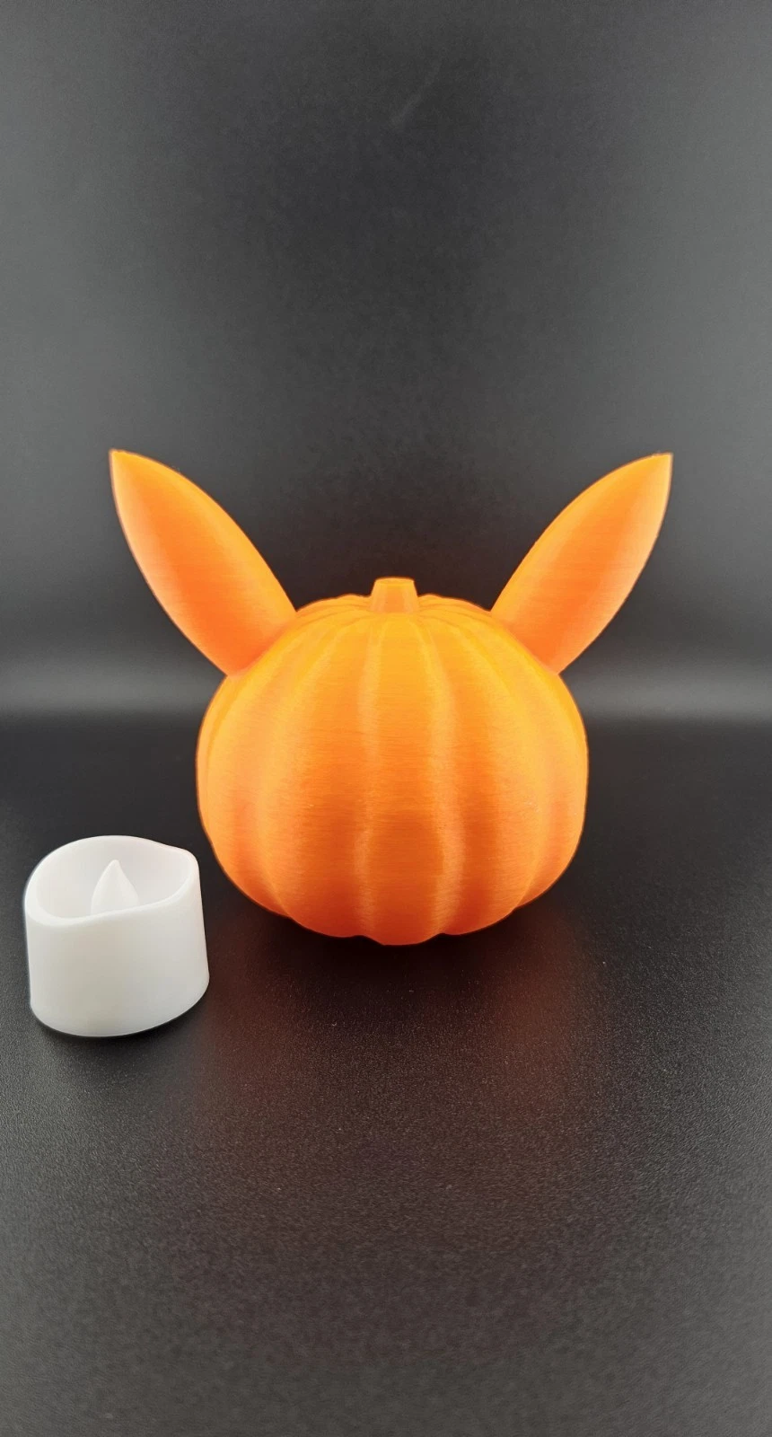 Orange 3D-printed pumpkin pikachu and a white tea light on a black surface.