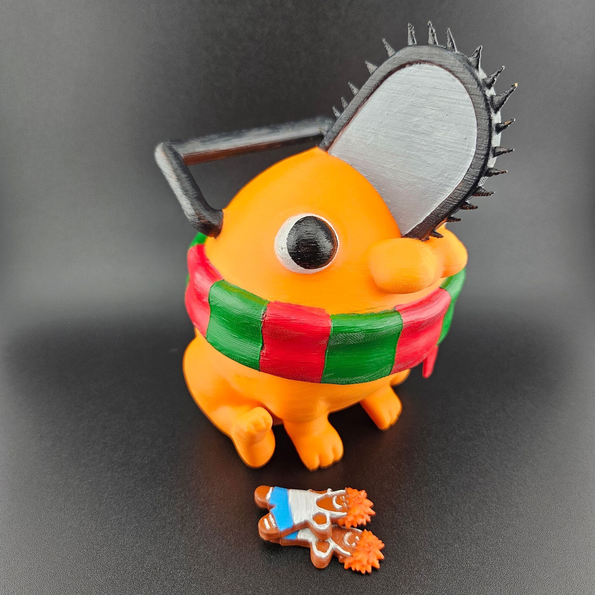Orange Pochita figurine with chainsaw head, wearing a Christmas scarf and holding gingerbread.
