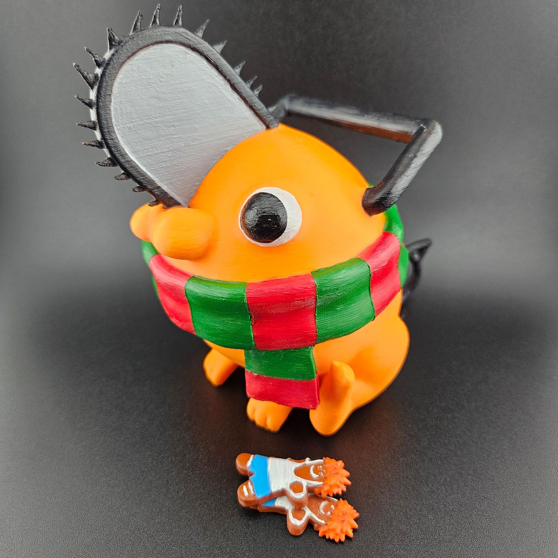 Orange Pochita figurine with chainsaw head, wearing a Christmas scarf and holding gingerbread.