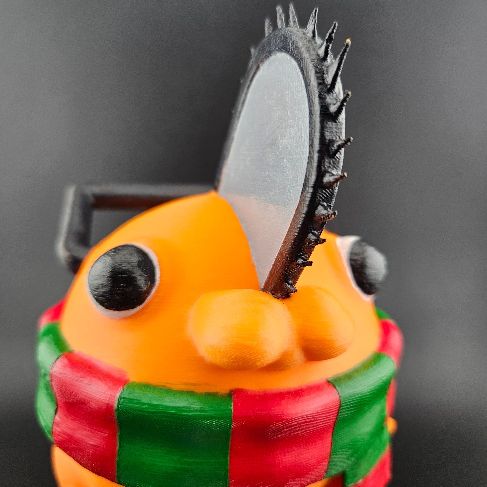 Orange figure with chainsaw head, black eyes, wearing a red and green scarf, against a dark background.