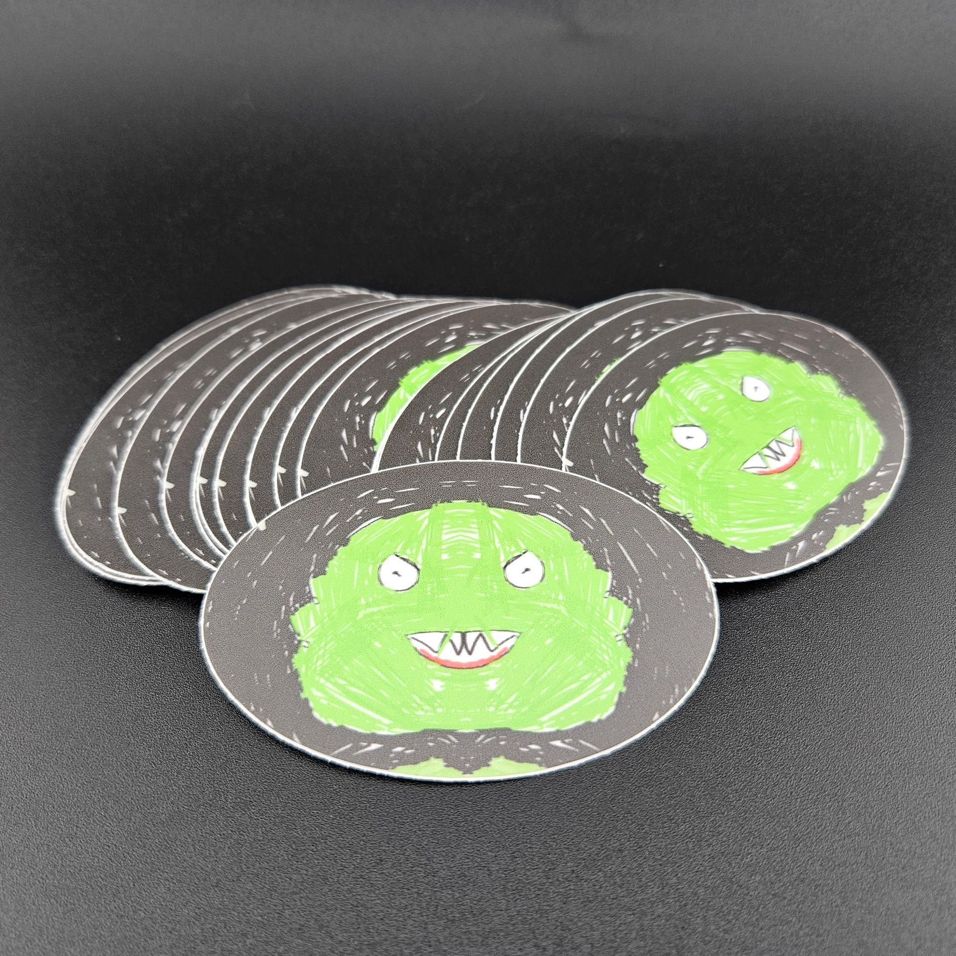 Stack of green monster-faced stickers with black borders, on a black surface.