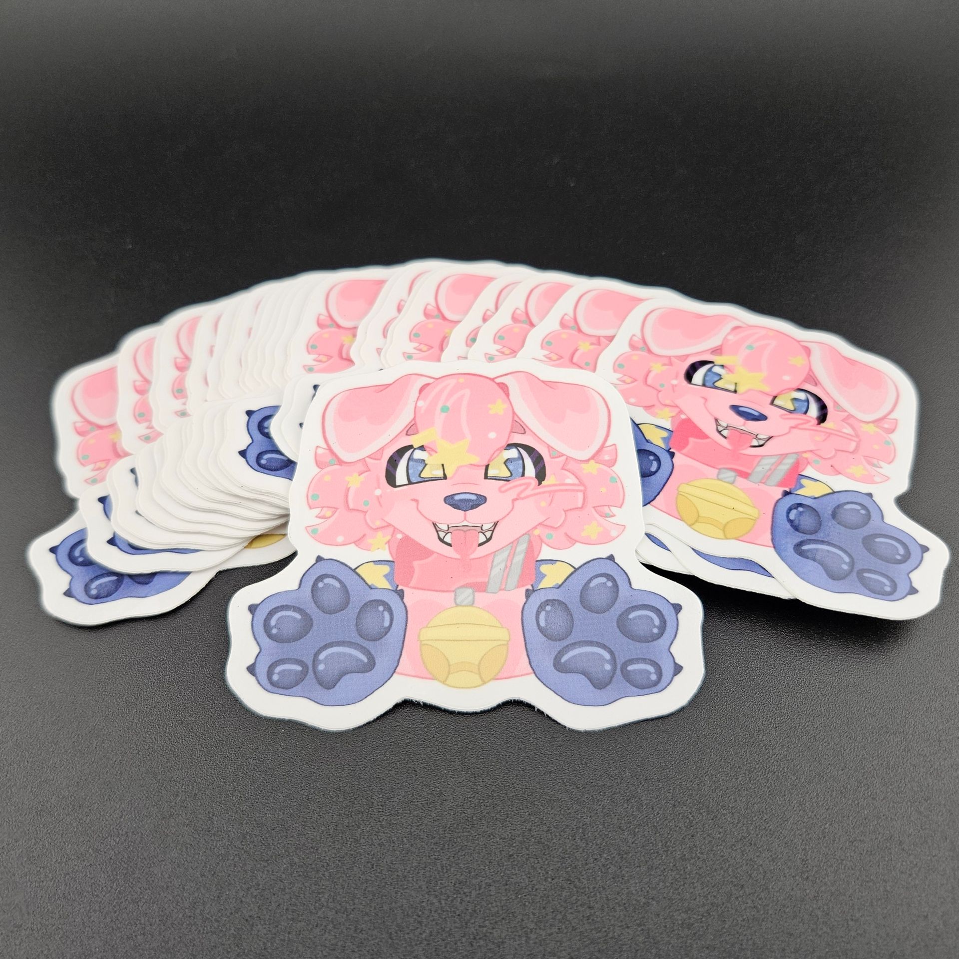 Pile of pink and blue cartoon dog stickers with open arms and friendly expression on black surface.