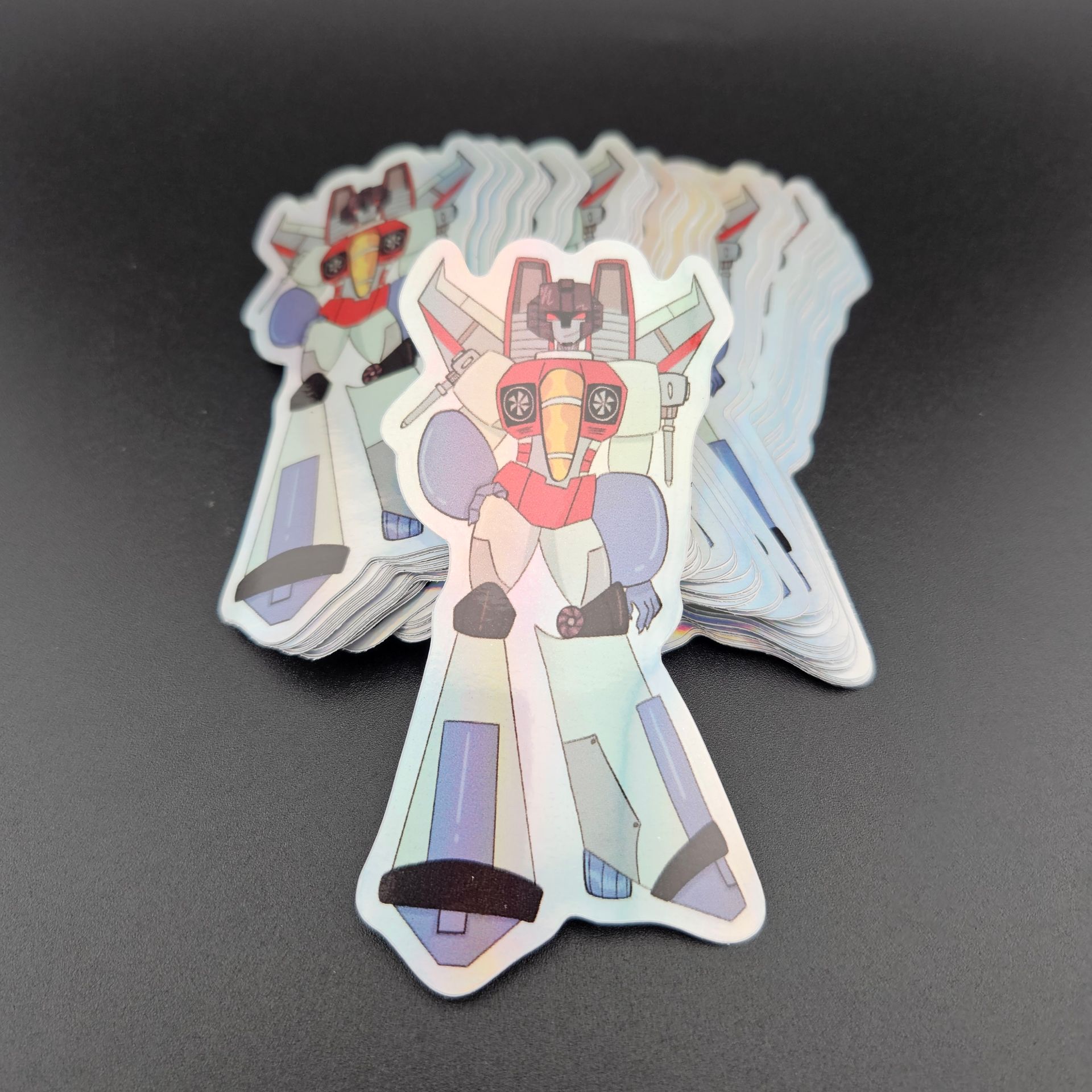 Holographic Starscream stickers stacked on a dark surface, robot cartoon figure with red, blue, and black details.