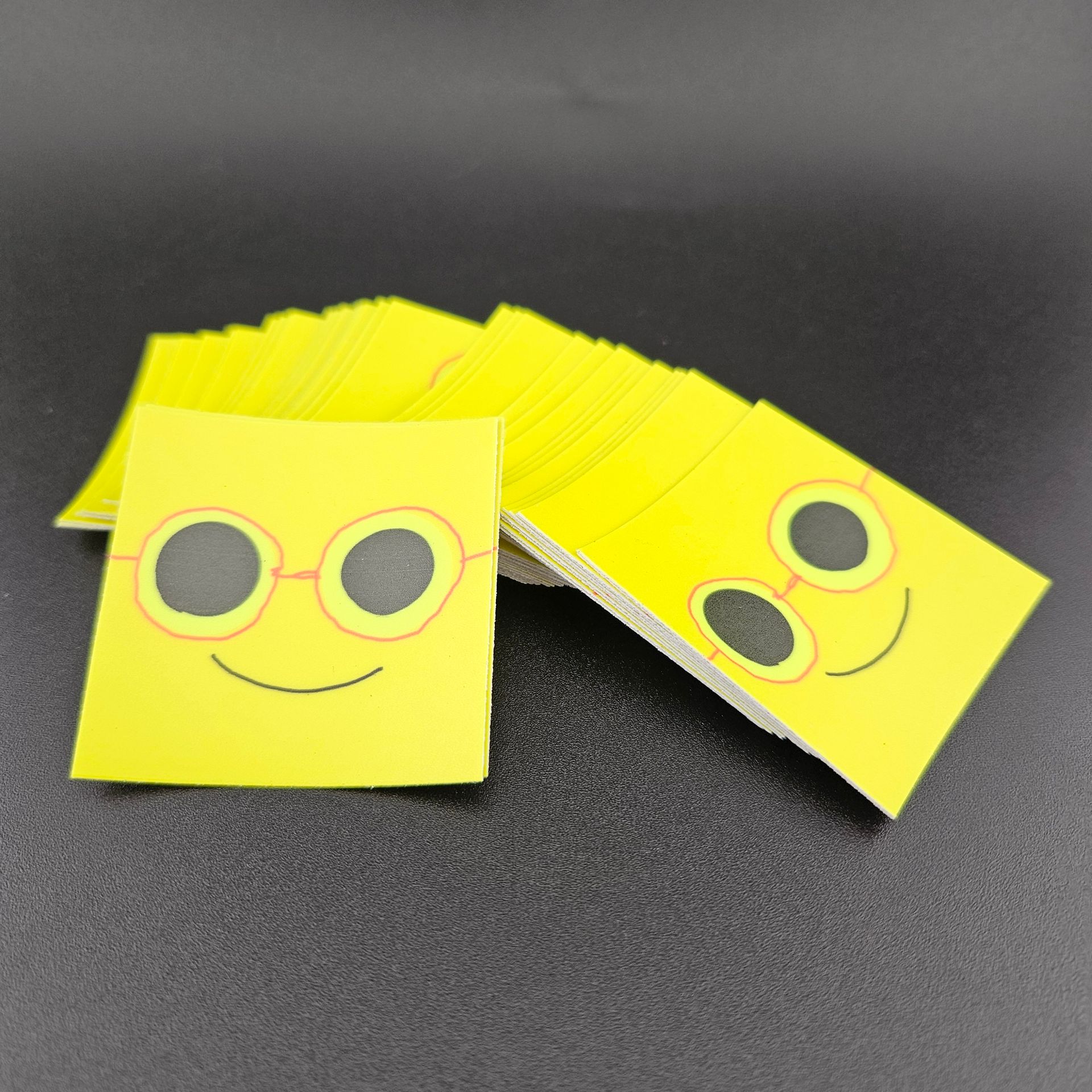 Yellow cards with smiling faces wearing glasses stacked on a dark surface.