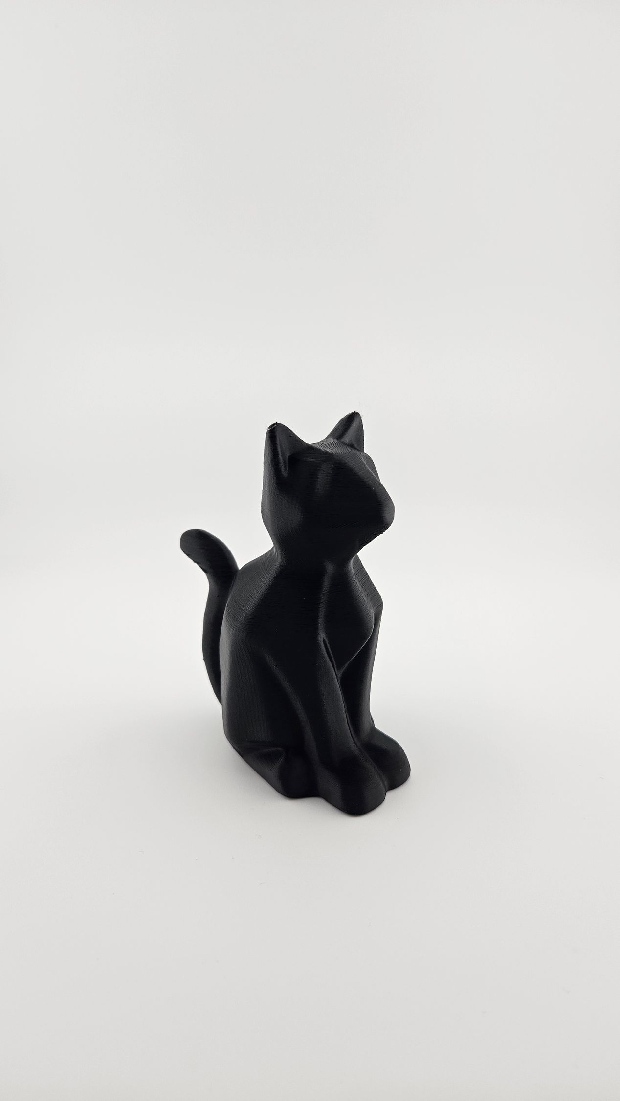 Black cat figurine, sitting upright, looking up, on a white surface.