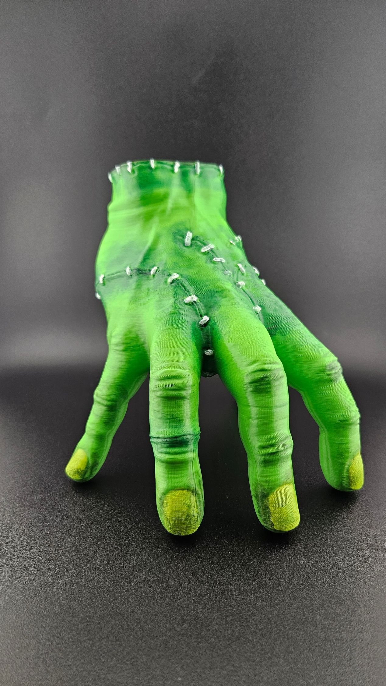 Green Frankenstein-like hand with stitches and metal studs, on a black background.