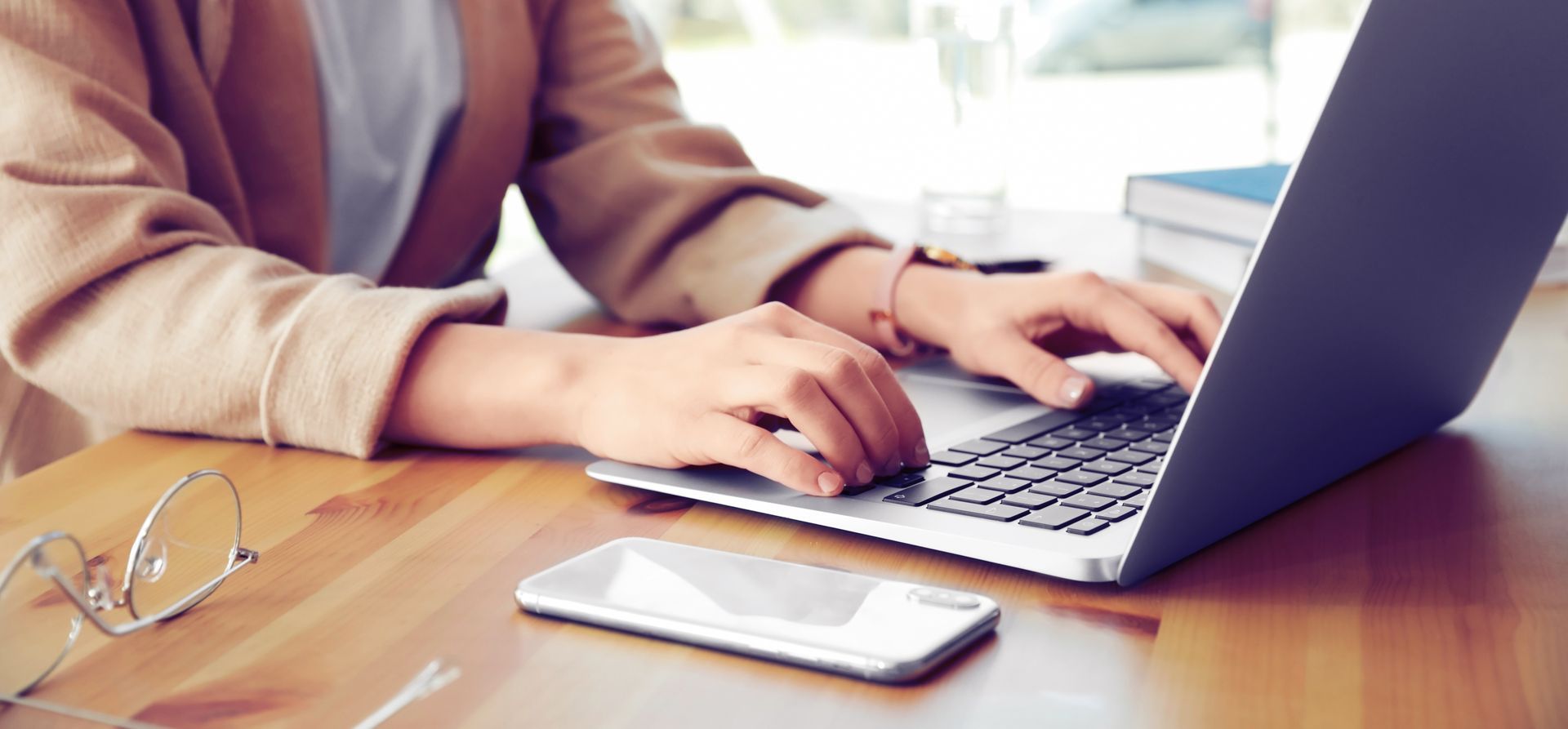 Woman typing on silver laptop in beige coat. Sat at desk with books and glasses beside her