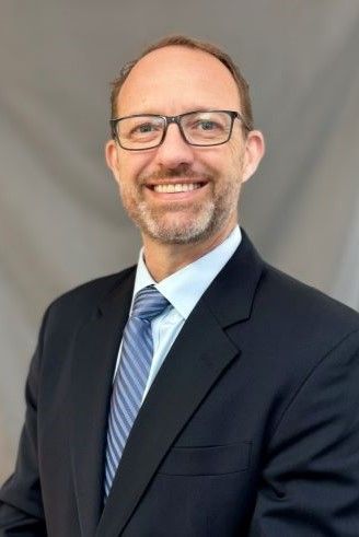 Man wearing glasses and a suit, smiling, with a light blue tie.