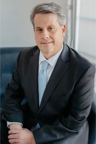 Man in a dark suit and light blue tie sitting on a dark chair, smiling.