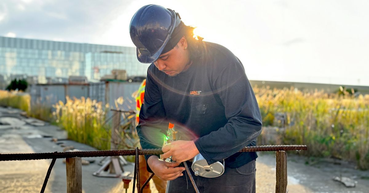Person in hard hat using a tool outdoors near a waterfront at sunset, with a bright sun flare.