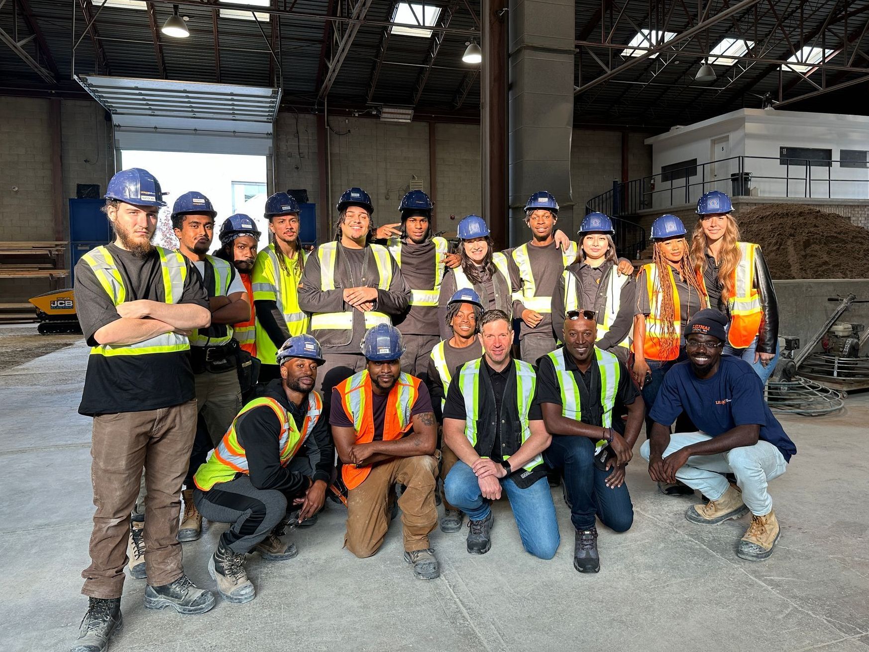 Mark and Joe sitting at a table in front of microphones. A group of construction workers are posing for a picture in a warehouse.