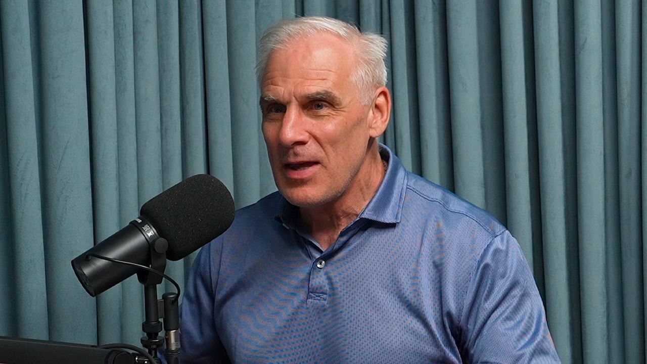 Man speaking into a microphone in front of blue curtains, wearing a blue shirt.