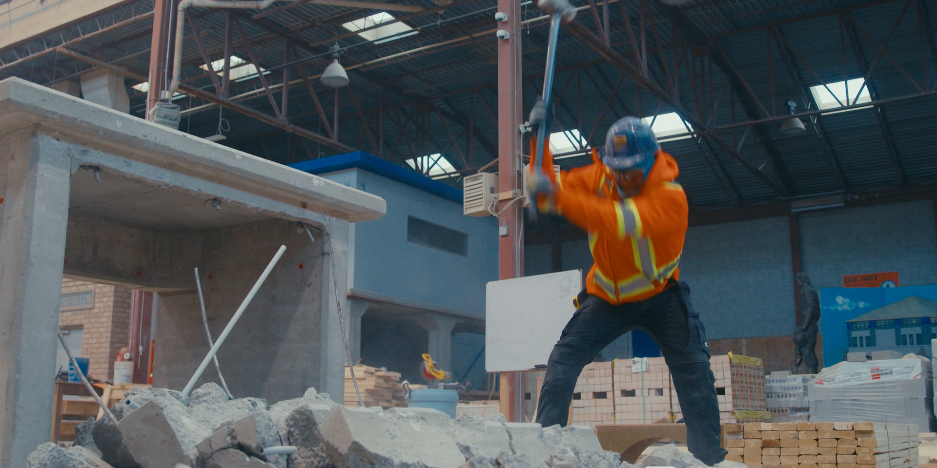 Construction worker in orange jacket and safety vest swings a sledgehammer at a pile of rubble.