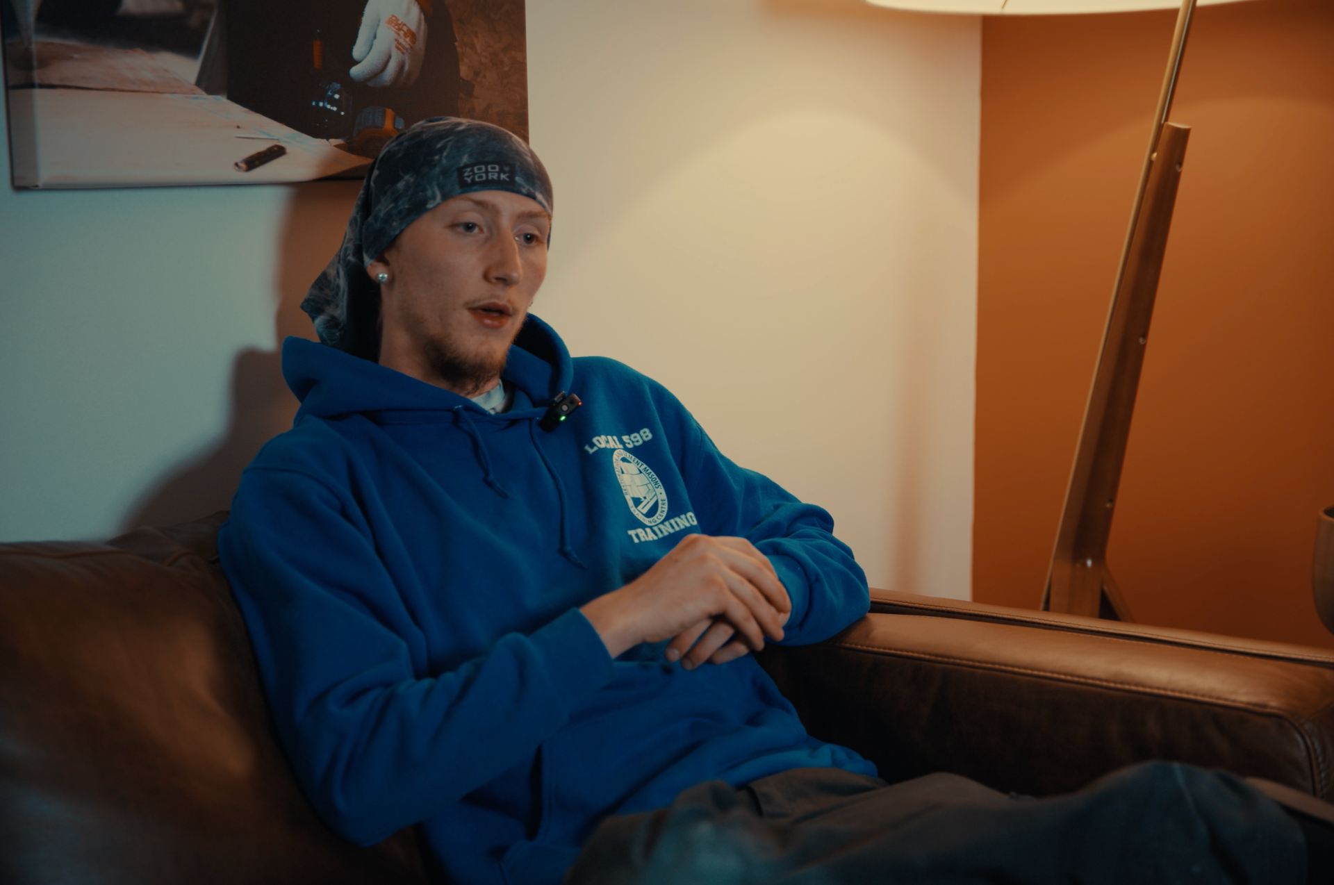 Person in a blue hoodie sits on a couch in a warmly lit room, looking thoughtful.