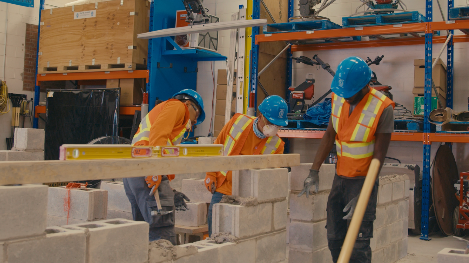 Construction workers building a cinder block wall, using a level. Indoor setting with equipment and storage.