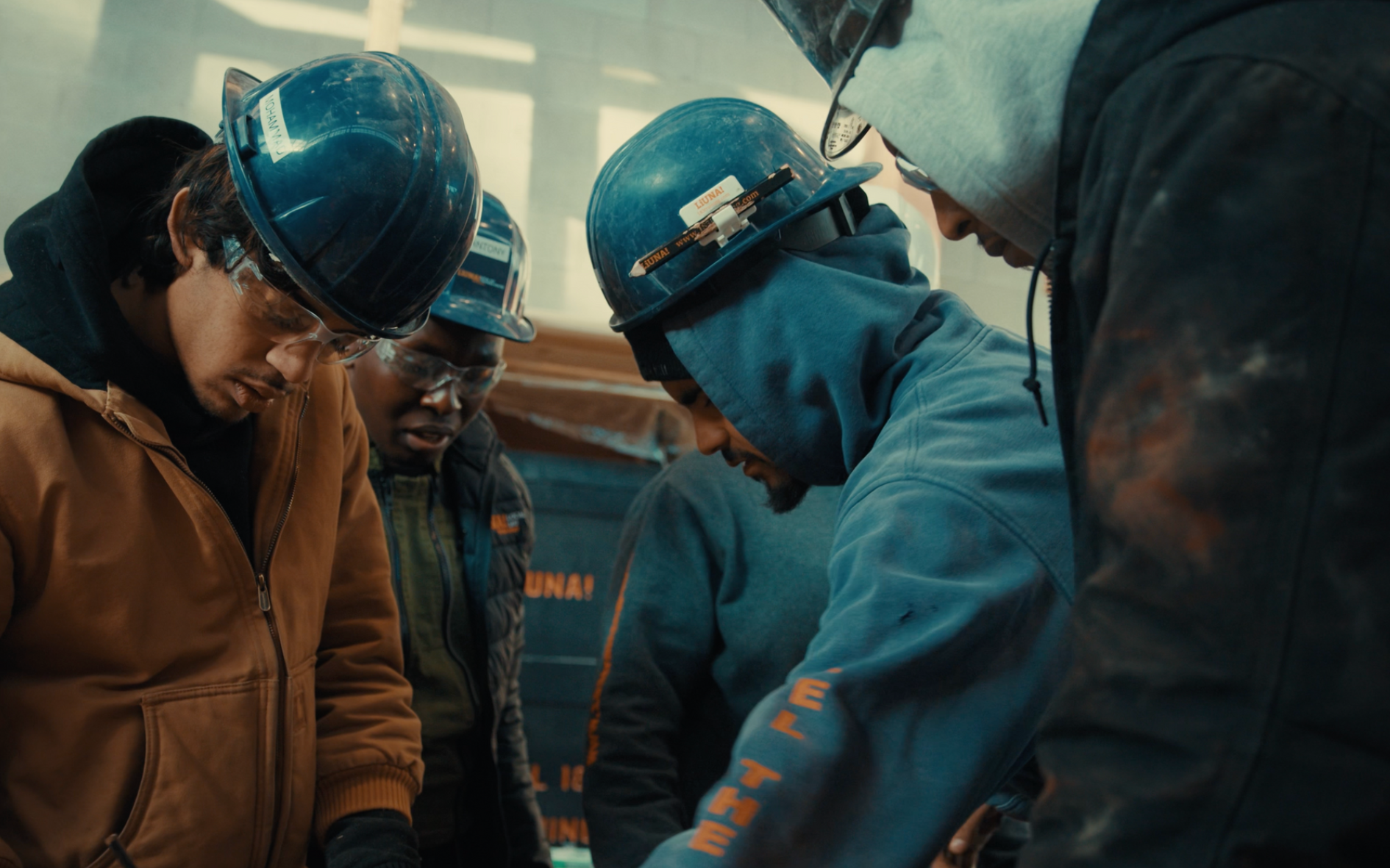 Workers in blue hard hats and jackets huddle around a task on a construction site.