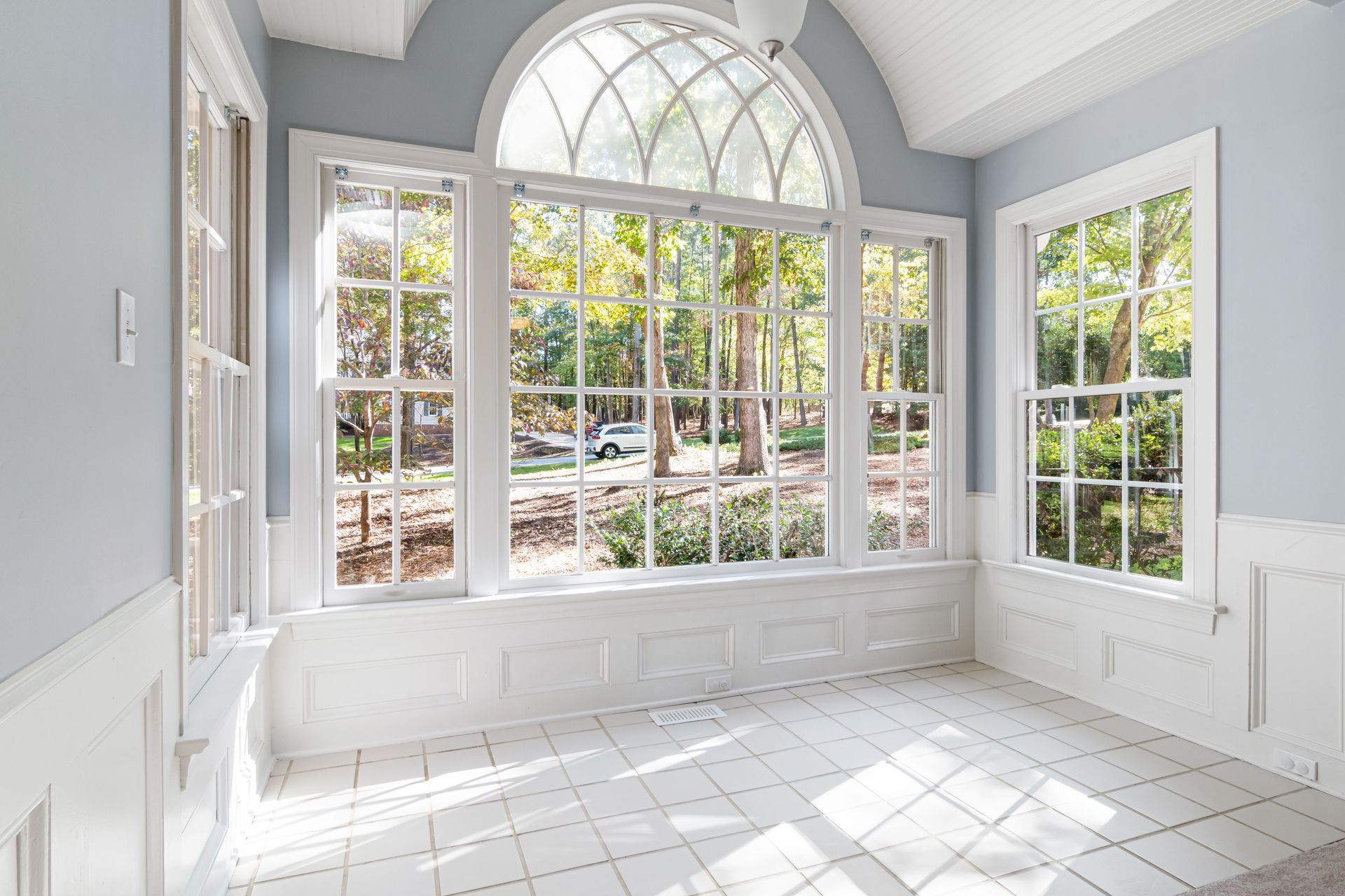 Bright sunroom with large arched windows, white tile floor, and pale blue walls overlooking a garden.