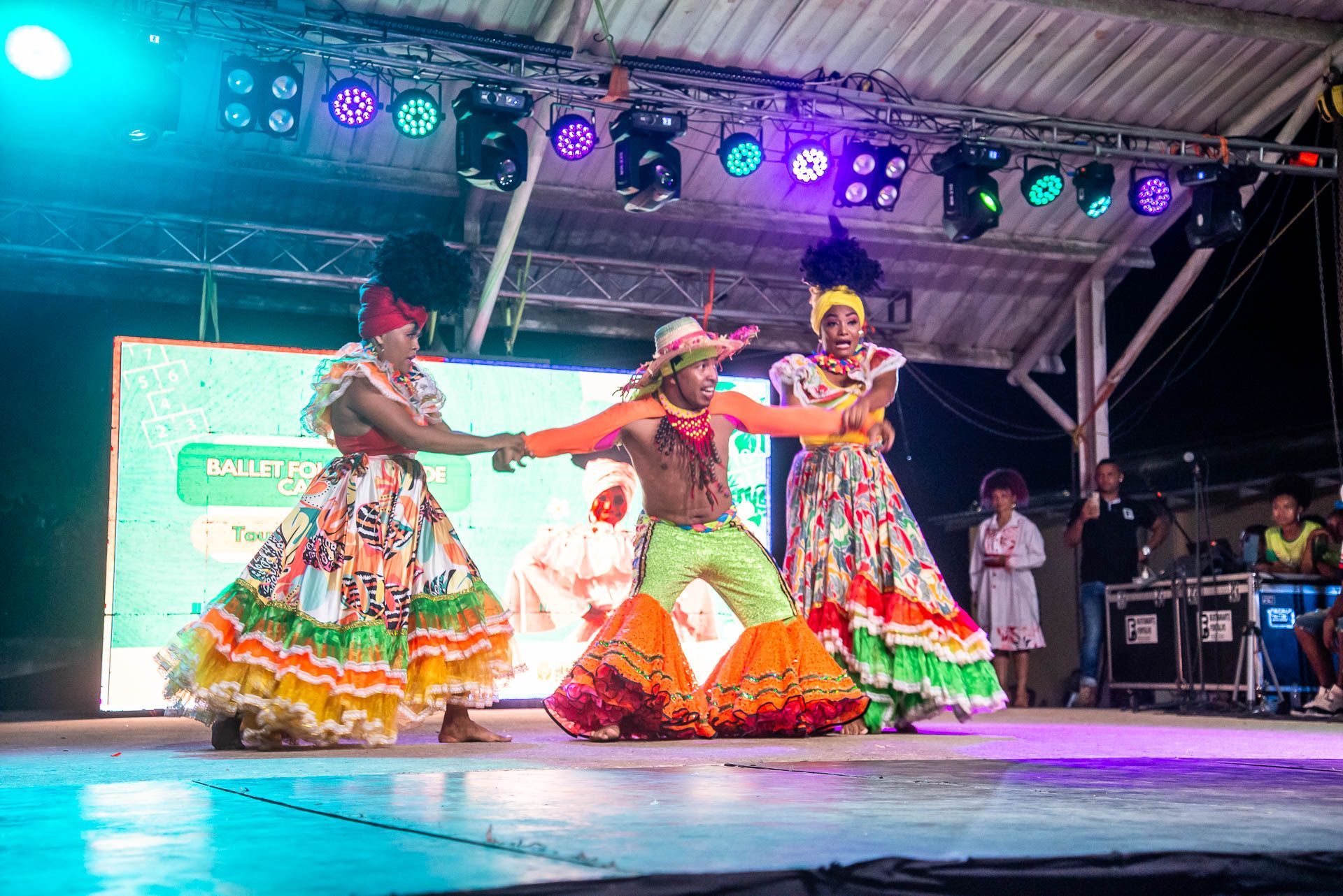 Tres artistas con trajes coloridos bailan en un escenario bajo las luces del escenario en un festival.